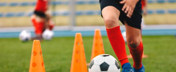 Footballer dribbling ball on training between orange cones. Young football player in sports blue cleats and red socks