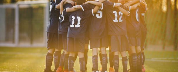 Group of children huddling with coach. Summer sunset at the stadium in the background. Youth soccer football team group photo. Happy boys soccer players kicking tournament. School boys in blue jerseys