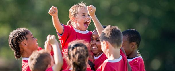 A multi-ethnic soccer team full of elementary age children are cheering together after winning their game. They have their arms raised up in the air victoriously.
